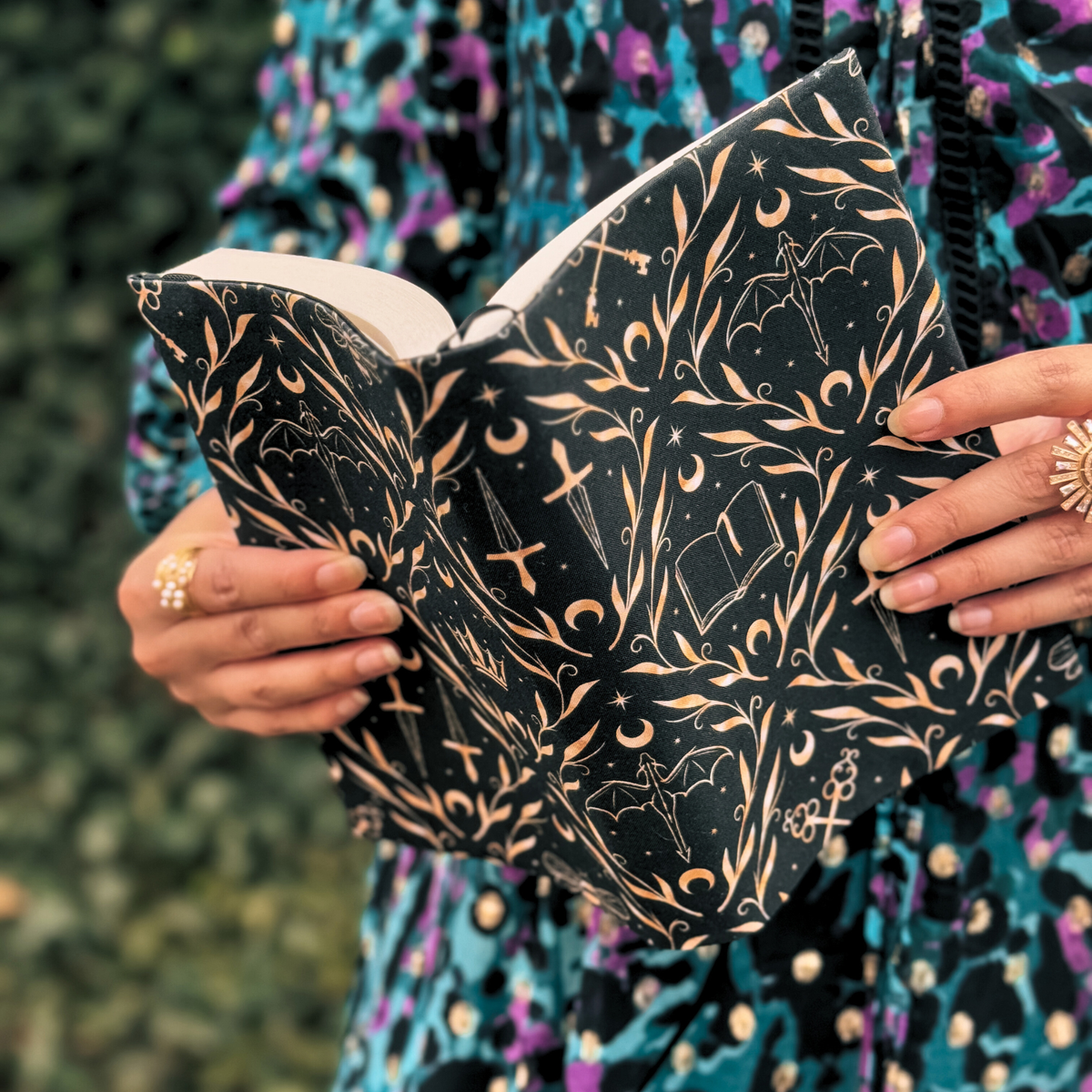 A women holding an open book with a black fabric dust jacket decorated with gold books, daggers, moons and stars against a blurred background.