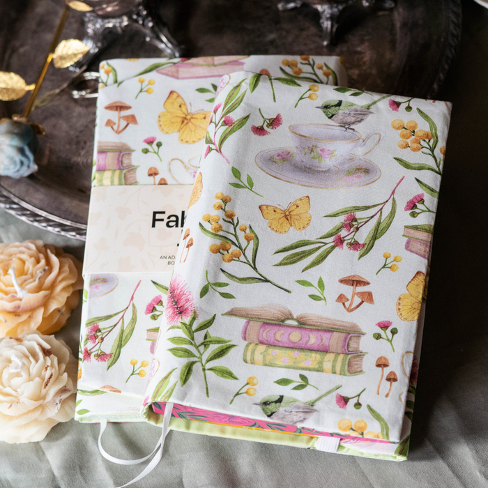 A book in a white fabric dust jacket decorated in pink and green plants, yellow butterflies, stacks of books, and mushrooms on top of a able decorated with yellow flowers. 
