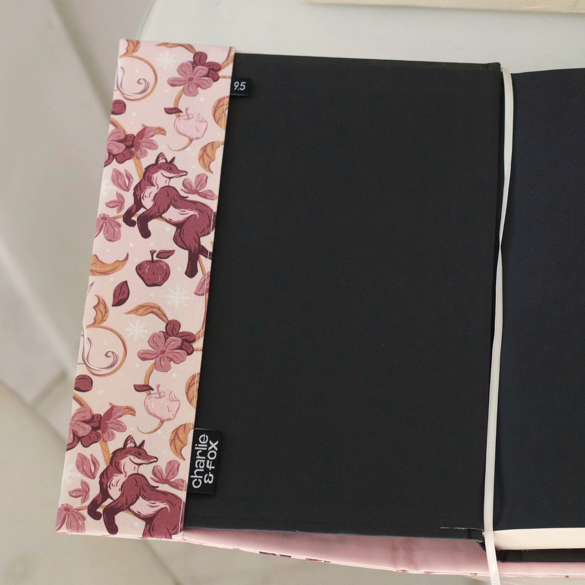 Close up of an open book with a light pink fabric dust jacket decorated with foxes, pink flowers light pink apples, bow and arrows, and gold swirls on top of a white table