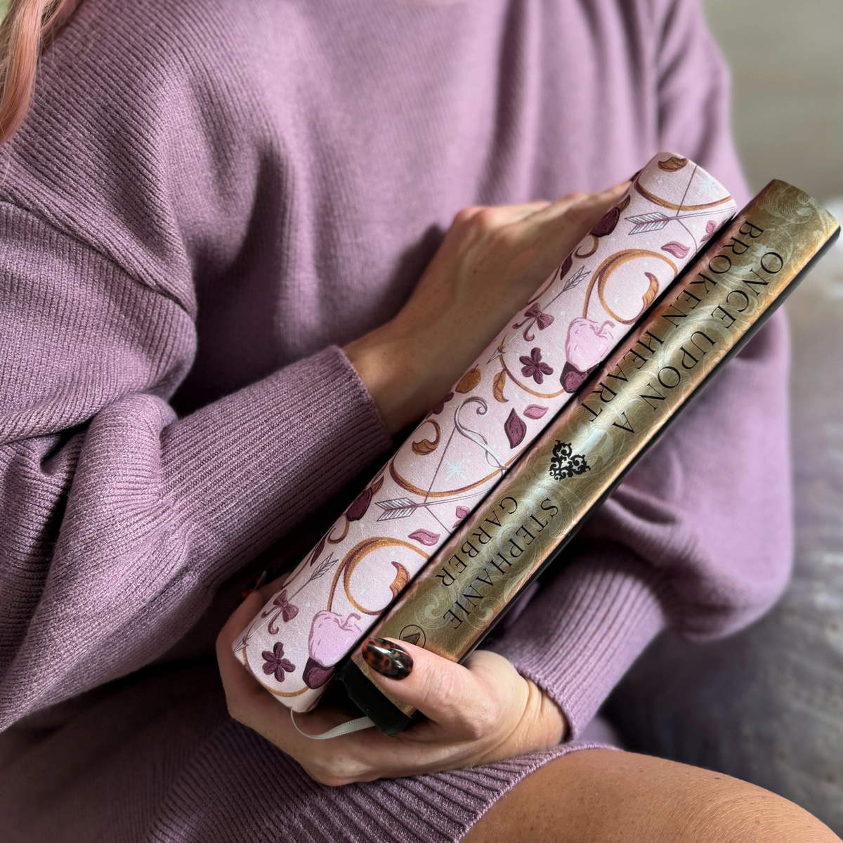 A women holding two books one with a light pink fabric dust jacket decorated with foxes, pink flowers light pink apples, bow and arrows, and gold swirls and the other titled Once Upon a Broken Heart sitting on a couch